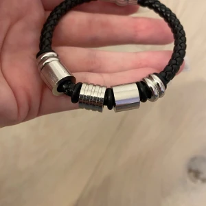 Svart flätat armband med silverdetaljer - Stilrent svart armband med flätad design och flera coola silverfärgade metallringar. Magnetlås i silverfärg gör det enkelt att ta på och av. Perfekt accessoar för en edgy look.