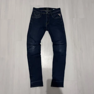 Replay Anbass jeans mörkblå 29/32 - Snygga Replay Anbass jeans i mörkblå tvätt med klassisk femficksdesign och kontrastsömmar. Modellen har en slim passform och raka ben, med Replay-logga på fickan. Perfekt för dig som gillar stilrena och moderna jeans.