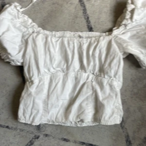 Vit blus med puffärmar och smock - Säljer en söt vit blus med puffiga korta ärmar och smockad rygg. Toppen har en fyrkantig urringning och resår vid ärmslut och nederkant för en bekväm passform. Perfekt för dig som gillar romantisk och trendig stil.
