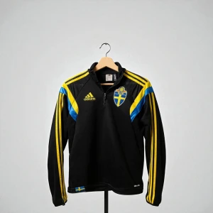 Svenska landslaget långärmad tröja Adidas - Snygg svart långärmad fotbollströja från Adidas med Sveriges landslagsemblem på bröstet. Tröjan har gula och blå detaljer, dragkedja vid halsen och klassiska Adidas-ränder längs ärmarna. Perfekt för dig som vill visa ditt stöd för Sverige.