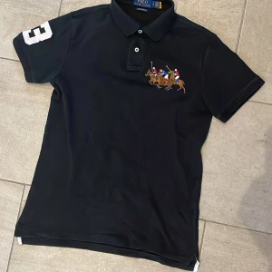 Svart pikétröja Polo Ralph Lauren S - Svart pikétröja från Polo Ralph Lauren i custom slim fit. Klassisk krage med knappar och broderat polospel-motiv på bröstet. Stor vit siffra 3 på ärmen. Tillverkad i bomull, perfekt för en sportig och stilren look.