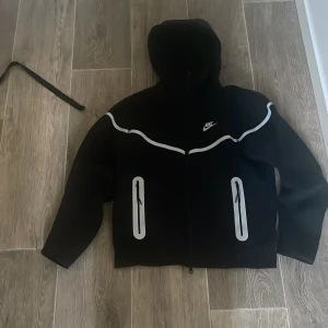 Nike tech fleece byxor och kofta - Svart vindjacka från Nike med huva och vita reflexdetaljer längs bröstet och vid fickorna. Jackan har dragkedja framtill och två vertikala fickor med dragkedja. Perfekt för dig som vill sticka ut med snygga reflexer när det är mörkt ute. M men passar exakt som S 