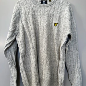  Lyle & Scott - Säljer en grå kabelstickad tröja från Lyle & Scott med deras klassiska gula logga på bröstet. Tröjan har rund halsringning, långa ärmar och ribbade muddar. Perfekt för lager-på-lager och chill stil. Storlek L, men mindre i storlek