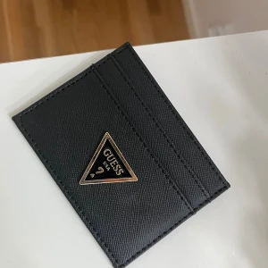 Svart kortfodral från Guess - Snyggt svart kortfodral från Guess med klassisk trekantslogga i guld och svart framtill. Fodralet har flera kortfack och är gjort i ett strukturerat skinnmaterial som känns både stilrent och modernt. Perfekt för dig som vill ha koll på korten utan att ta plats.