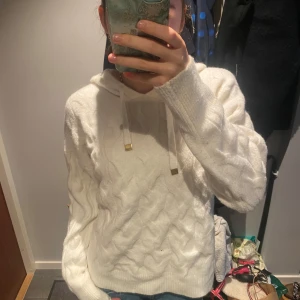 Vit stickad hoodie med snörning - Mysig vit stickad hoodie med kabelstickat mönster och snörning i huvan. Tröjan har långa ärmar och ribbade muddar vid ärmslut och nederkant. Perfekt för kyliga dagar och enkel att matcha med jeans eller leggings.