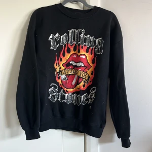 Rolling Stones svart sweatshirt - Svart sweatshirt med stort Rolling Stones-tryck framtill, eldflammor och klassiska röda tungan. Tröjan har rund hals, långa ärmar och ribbade muddar. Perfekt för dig som gillar bandmerch och vill sticka ut med en ikonisk look.