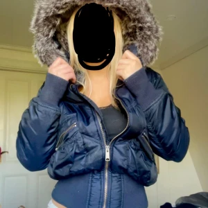 Mörkblå pufferjacka med fuskpäls - Säljer en mörkblå pufferjacka med fluffig fuskpäls på huvan från Oasis. Jackan har två stora fickor med dragkedja framtill och ribbade muddar vid midja och ärmslut. Croppad modell som ger en cool siluett, perfekt för kalla dagar.