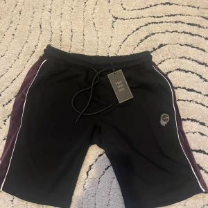 Svarta shorts från La Vichy XS - Snygga svarta shorts från La Vichy i storlek XS med lila sidopaneler i mjuk sammet och vita kantlinjer. De har snörning i midjan och ett broderat emblem på benet. Perfekta för chill eller träning, riktigt skön passform och cool design.