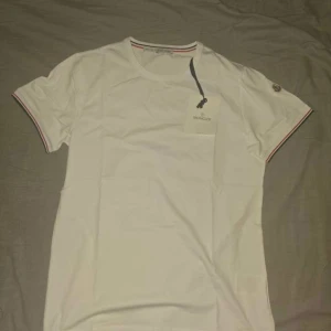 Vit slim fit t-shirt från Moncler - Vit t-shirt från Moncler i slim fit-modell. Klassisk rund halsringning och korta ärmar med röd, vit och blå rand med Moncler-logga på vänster ärm. Skön bomullskvalitet och stilren design som sticker ut med snygga detaljer. Denna tshirt har aldrig använts så den är i nyskick och har fortfarande taggarna på. Pris kan diskuteras! och kan mötas upp i Skåne.  