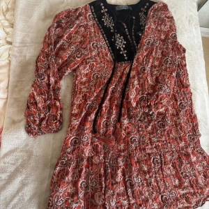 Mönstrad klänning från Primark - Boho-inspirerad kortklänning från Primark med röd, svart och vit paisleymönster. Klänningen har långärmade puffärmar, broderad svart sammet vid bysten, v-ringning och knytband framtill. Perfekt för dig som gillar unika detaljer och färgstarka plagg.