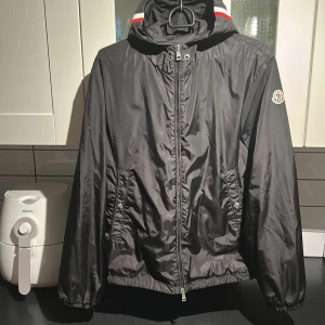 Svart vindjacka från Moncler - Svart vindjacka från Moncler med huva och coola rödvitabå detaljer på kragen. Jackan har dragkedja framtill, två fickor med tryckknappar och Moncler-logga på ärmen. Materialet är lätt och syntetiskt, perfekt för blåsiga dagar. 