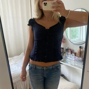 Mörkblå blus med volangdetaljer - Säljer en söt mörkblå blus med knappar framtill, volangkant vid axlar och nederkant samt liten rosettdetalj på axlarna. Blusen har v-ringning och är ärmlös, perfekt för en trendig och feminin look.