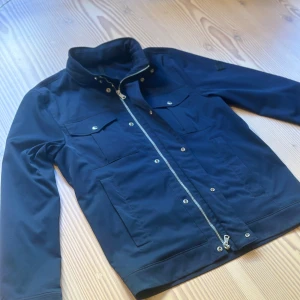 Mörkblå windbreaker från J.Lindeberg - Snygg mörkblå vindjacka från J.Lindeberg med utfällbar luva, flera fickor med tryckknappar och dragkedja framtill. Jackan har diskret logga på ärmen och är tillverkad i ett slätt, tåligt material. Inga defekter endast lite tecken på användning 