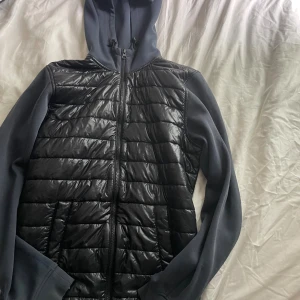 Svart pufferjacka Peak Performance S - Snygg svart pufferjacka från Peak Performance i storlek S. Jackan har huva med snörning, quiltad framsida och slät baksida. Dragkedja hela vägen och långa ärmar. Perfekt för dig som gillar sportig och clean stil.