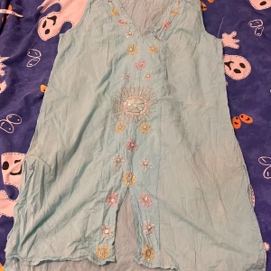 Ljusblå ärmlös klänning med broderi - Supersöt ljusblå klänning med v-ringning och broderade blommor i pastellfärger framtill. Klänningen är ärmlös, har slits framtill och är i ett lätt, luftigt material. Perfekt för varma dagar och ger en bohemisk vibe.