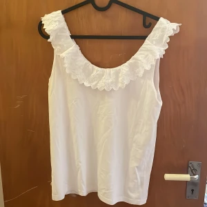 Vit blus med spetsvolang. - Söt, vit blus med bred, broderad spetsvolang runt urringningen. Ärmlös modell i mjukt bomullstyg. Osäker på märke men det finns en rund platta i sidan på blusen med något tecken❣️