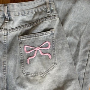 Ljusblåa jeans med rosa rosetter - Snygga grå jeans med raka ben och broderade rosa rosetter på bakfickorna. Den söta detaljen eleverar varje look.🎀