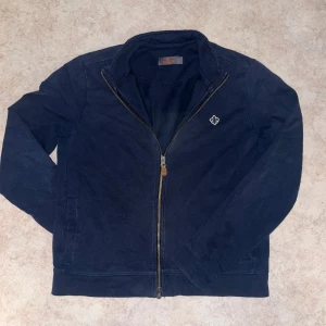 Mörkblå tjock tröja från Morris - Snygg mörkblå tjocktröja från Morris med dragkedja framtill och diskret logga på bröstet. Tjocktröjan har en klassisk krage och raka ärmar Enkel och stilren design som funkar till det mesta.