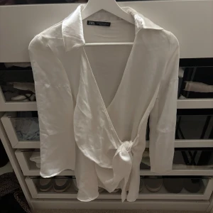 Vit omlottblus klänningen från Zara i sidenlook - Säljer en vit omlottblus från Zara med snygg v-ringning och knyt i midjan. Blusen har trekvartsärm och är gjord i ett glansigt material som ger en lyxig känsla. Perfekt för dig som gillar stilrena och trendiga plagg.