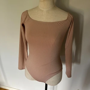 Ribbad beige bodysuit från NLY One - Säljer en ribbad beige bodysuit från NLY One i storlek S. Den har lång ärm, bred urringning och är figurnära i modellen. Perfekt att styla med jeans eller kjol för en clean look. Materialet är stretchigt och mjukt för extra komfort.
