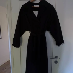 Svart lång trenchcoat från Wera - Stilren svart lång trenchcoat från Wera med markerad krage, knytskärp i midjan och långa ärmar. Trenchcoaten har en klassisk omlott-look och dolda knappar framtill. Perfekt för dig som gillar minimalistisk och tidlös stil.