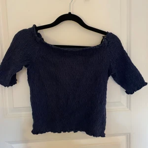 Mörkblå offshoulder topp - Säljer en mörkblå offshoulder topp med smockad struktur och korta ärmar. Toppen har en tight passform och vågig kant nertill och vid ärmsluten. 