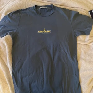 Blå Stone Island t-shirt - Snygg blå t-shirt från Stone Island med klassisk logga i gult och vitt på bröstet. T-shirten har rund hals och korta ärmar, perfekt för en avslappnad och stilren look. Tillverkad i mjuk bomull som känns skön mot huden.nypris 1100. Frakt står köparen för 