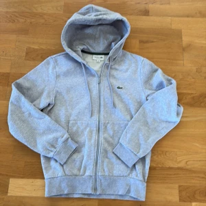 Grå hoodie från Lacoste med dragkedja - Snygg ljusgrå hoodie från Lacoste i klassisk passform. Tröjan har huva med snörning, dragkedja framtill och den ikoniska krokodilloggan på bröstet. Tillverkad i mjuk bomullsmix, perfekt för chill dagar eller när du vill vara casual. Hann inte använda så många gånger innan jag växte ur den.