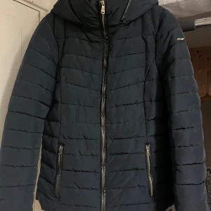 Blå pufferjacka med huva - Snygg blå  pufferjacka med huva och dragkedja framtill. Från Esprit .Jackan har två fickor med dragkedja och horisontella sömmar som ger en quiltad look.väldigt varm så  Perfekt för kyliga dagar och enkel att matcha med olika outfits.