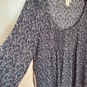 Blåmönstrad blus från 365 Sunshine - Supersöt småblommig blus från 365 Sunshine i storlek 36. Blusen har små knappar framtill, rund halsringning och trekvartsärm. Mönstret är i blått, lila, rosa, och svart på ljus botten. Lätt transparent och kan alternativt knytas i midjan, tillhörande litet skärp medföljer.