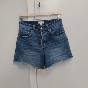 Högmidjade jeansshorts från H&M - Snygga blå jeansshorts från H&M i klassisk modell med hög midja och råa, fransiga benslut. Shortsen har fem fickor, knappgylf och en avslappnad passform. Perfekta för sommardagar och festivalhäng.