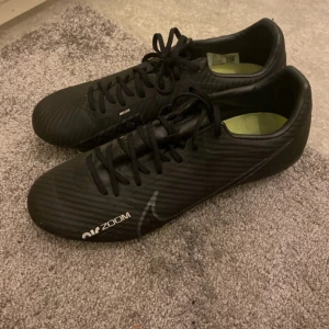 Nike Mercurial Air Zoom fotbollsskor - Säljer ett par svarta Nike Mercurial Air Zoom fotbollsskor med snörning och diskret mönstrad ovandel. Skorna har en smal och sportig form, med vita detaljer och text på sidan och hälen. Perfekta för dig som vill ha grymt grepp och stil på planen.