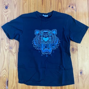 Svart Kenzo t-shirt  - Svart t-shirt från Kenzo med ikoniskt blått tigertryck och texten 'Kenzo Paris' framtill. Klassisk rund halsringning och korta ärmar. Perfekt för dig som vill sticka ut med en streetig vibe och cool design.