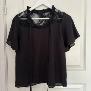 Svart blus med spets från SHEIN - Svart blus från SHEIN med transparent spets upptill och volangdetaljer vid kragen. Knytband framtill och korta, fladdriga ärmar ger en romantisk vibe. Perfekt för dig som gillar detaljer och en lite dressad look.