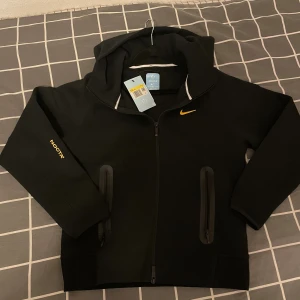 Svart NOCTA x Nike hoodiejacka xs - Svart hoodiejacka från Nike NOCTA med gul logga på bröstet och NOCTA-tryck på ärmen. Jackan har dragkedja framtill, två fickor med dragkedja och en sportig vibe. Perfekt för dig som gillar streetwear och snygga detaljer.