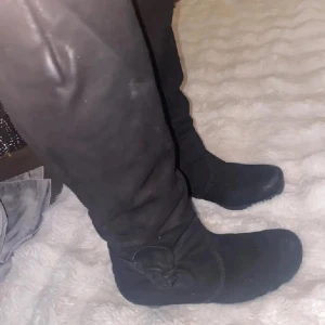 Svarta mockastövlar med klack - Snygga svarta stövlar i mocka med dragkedja på sidan och låg klack. Stövlarna har en rund tå och en dekorativ rosett på utsidan. Perfekta för höst och vinter när du vill ha en stilren look.