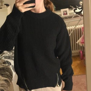 Svart stickad tröja med dragkedjor - Säljer en svart, grovstickad tröja med lång ärm och rund hals. Tröjan har coola dragkedjor i sidorna nertill som ger en extra edge. Passformen är loose och den är perfekt för lager på lager under höst och vinter.