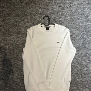 BOSS vit sweatshirt  - Säljer en stilren vit långärmad tröja från BOSS med diskret logga på bröstet och nacken. Tröjan har rund halsringning och är tillverkad i mjuk bomull, perfekt för en clean och enkel look.