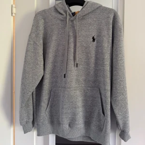 Grå hoodie från Polo Ralph Lauren - Helt oanvänd tunn grå hoodie från Polo Ralph Lauren med svart broderad logga på bröstet. Tröjan har huva med snörning, känguruficka och ribbade muddar. Perfekt för chill dagar och enkel att matcha med jeans eller joggers. Storlek L men skulle säga att den sitter som storlek M. OBS! Äkthet kan inte garanteras på denna! Modellen är 178 och väger 74 kg. 