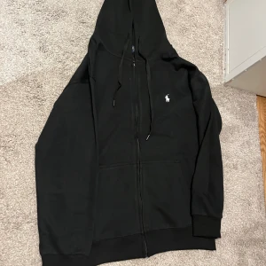 Svart hoodie från Polo Ralph Lauren - Svart hoodie från Polo Ralph Lauren med klassisk vit logga på bröstet. Tröjan har huva med snörning, dragkedja framtill och två fickor. Tillverkad i mjuk bomull, perfekt för chill dagar. Passformen är loose och ärmarna är långärmade.