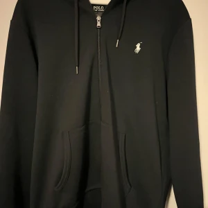Mörkblå hoodie från Polo Ralph Lauren - Mörk blå hoodie från Polo Ralph Lauren med vit broderad logga på bröstet. Tröjan har huva med snörning, magficka och half zip-dragkedja framtill. Perfekt för dig som gillar stilren och klassisk streetwear.