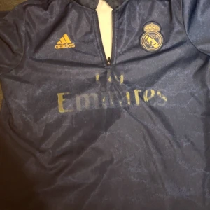 Real Madrid långärmad träningströja Adidas - Mörkblå långärmad Real Madrid träningströja från Adidas med gul logga och klubbmärke på bröstet. Tröjan har halv dragkedja framtill och är gjord i 100% polyester. Tunn gul detalj längs kragen och tryckt sponsor på magen.