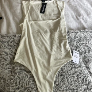 Vit ärmlös bodysuit från Boohoo - Snygg vit bodysuit från Boohoo med fyrkantig ringning och dubbla lager för en slät look. Ärmlös modell i stretchigt, slinky material som sitter tight och ger en clean vibe. Perfekt att styla med jeans eller kjol för en trendig outfit. Aldrig använd