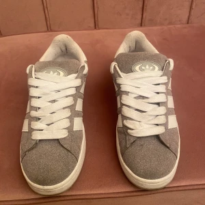 Grå Adidas Campus sneakers i mocka - Snygga Adidas Campus sneakers i grå mocka med vita detaljer och klassiska tre ränder på sidorna. Skorna har vit sula, rund tå och vita skosnören. Perfekta för dig som gillar en clean och tidlös streetstil.