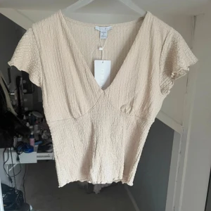 Beige omlottopp från MISU - Supersöt beige topp från MISU med omlott-look och v-ringning. Toppen har korta, lite fladdriga ärmar och är gjord i ett krinklat, lätt material som känns luftigt. Perfekt för varma dagar och enkel att styla med jeans eller kjol.