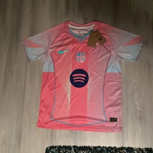 Barcelona  fotbollströja Nike - Säljer en Barcelona fotbollströja från Nike i en snygg rosa färg med ljusblå detaljer. Tröjan har korta ärmar, klubbmärke och sponsortryck på bröstet. Materialet är lätt och andas, perfekt för träning eller match. Modern design med grafiska linjer och mönster. Aldrig använt 