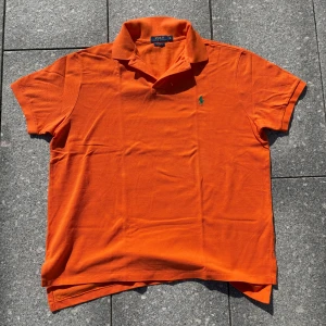 Orange pikétröja från Polo Ralph Lauren - Snygg orange pikétröja från Polo Ralph Lauren med klassisk krage och korta ärmar. Tröjan har det ikoniska gröna Polo-logot på bröstet och är gjord i mjuk bomull som andas. Perfekt för dig som gillar färg och vill sticka ut lite extra.