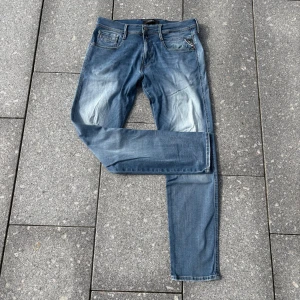 Replay Anbass Hyperflex jeans blå - Säljer ett par Replay Anbass Hyperflex jeans i ljusblå tvätt med slitningar och en trasig detalj på ena benet. Klassisk femficksmodell med stretchigt material och snygg faded effekt. Perfekta för dig som gillar en avslappnad och trendig look.