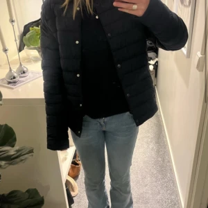 Svart pufferjacka från H&M - Säljer en svart pufferjacka från H&M i storlek 170. Jackan har en stilren look med rund halsringning, tryckknappar framtill och horisontella sömmar. Perfekt för kyliga dagar och enkel att matcha med olika outfits.