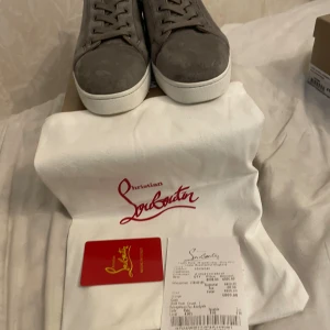 Gråa sneakers från Christian Louboutin - Snygga gråa sneakers från Christian Louboutin i mocka med vit platt sula och klassisk snörning. Skorna har en stilren och minimalistisk design som passar perfekt till streetwear eller en mer dressad look. Kommer med dustbag och äkthetskort.